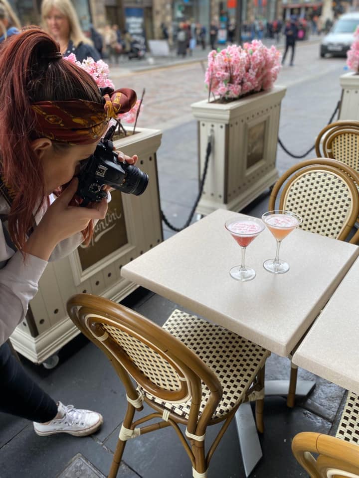 a person takes a photograph with a dslr camera of two cocktails in an outdoor seating area, surrounded by barriers with cherry blossoms on top